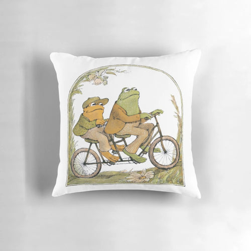 A charming pillow featuring two friends, a frog and a toad, biking together in a whimsical, cottagecore-inspired design.