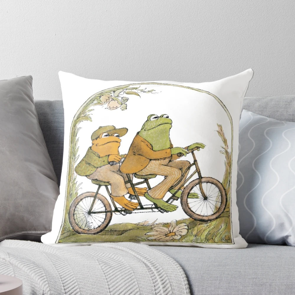 A charming pillow featuring two friends, a frog and a toad, biking together in a whimsical, cottagecore-inspired design.