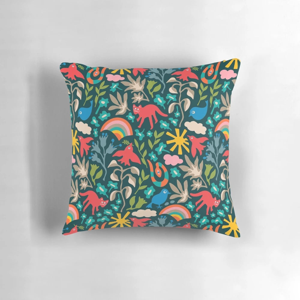 A colorful, nature-themed decorative pillow featuring birds, animals, flowers, rainbows, and clouds on a dark teal background.